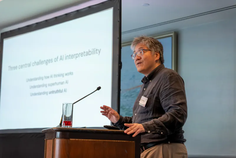 A speaker at the podium presenting a slide titled “Three central challenges of AI interpretability”