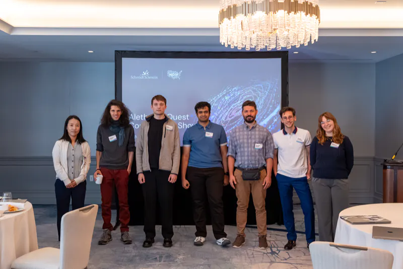 Seven blue team representatives standing in front of the Aletheia’s Quest Competition Showcase title slide: from left, Haein Kong of team ru, Tommaso Tosato of team Tara Research, Serhii Honcharenko of team FlaiChi, Jeremias Ferrao of team SAIN Groningen, Giuseppe Birardi of team EleutherAI, and Jacopo Zacchigna and Sara Candussio of team JSR